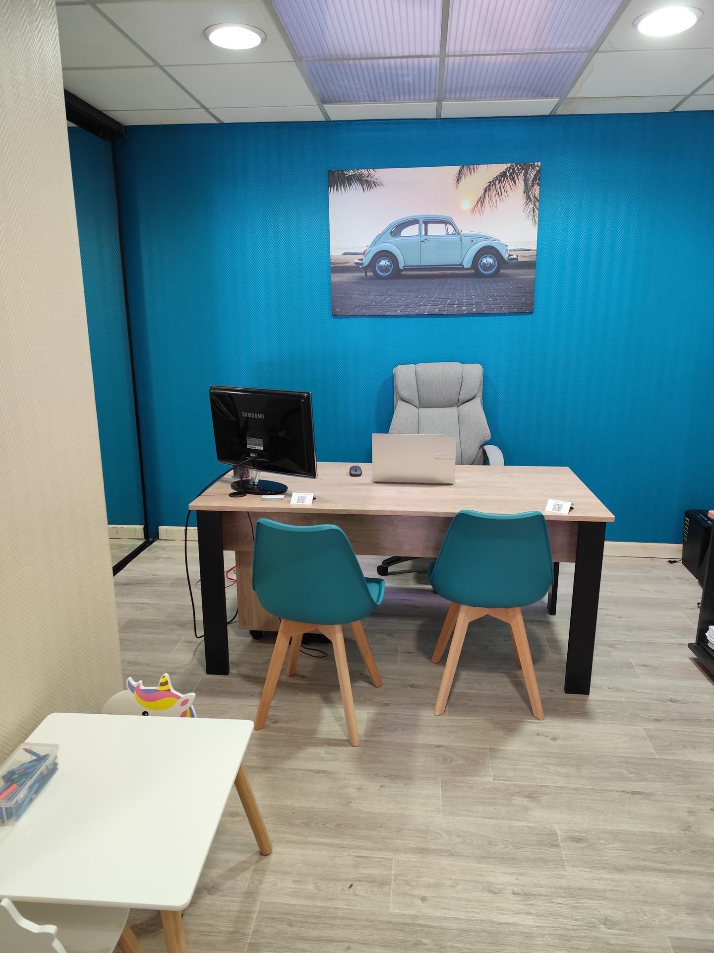 Petit bureau avec un mur turquoise, un bureau, un ordinateur et deux chaises bleu sarcelle sous une affiche de voiture.
