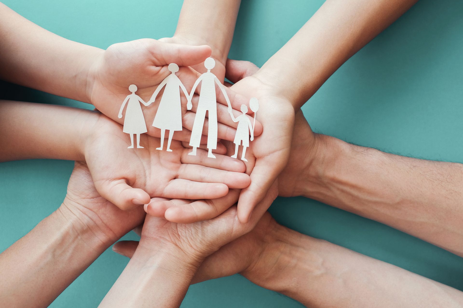 Mains de différentes personnes tenant des silhouettes familiales découpées dans du papier blanc sur un fond turquoise.