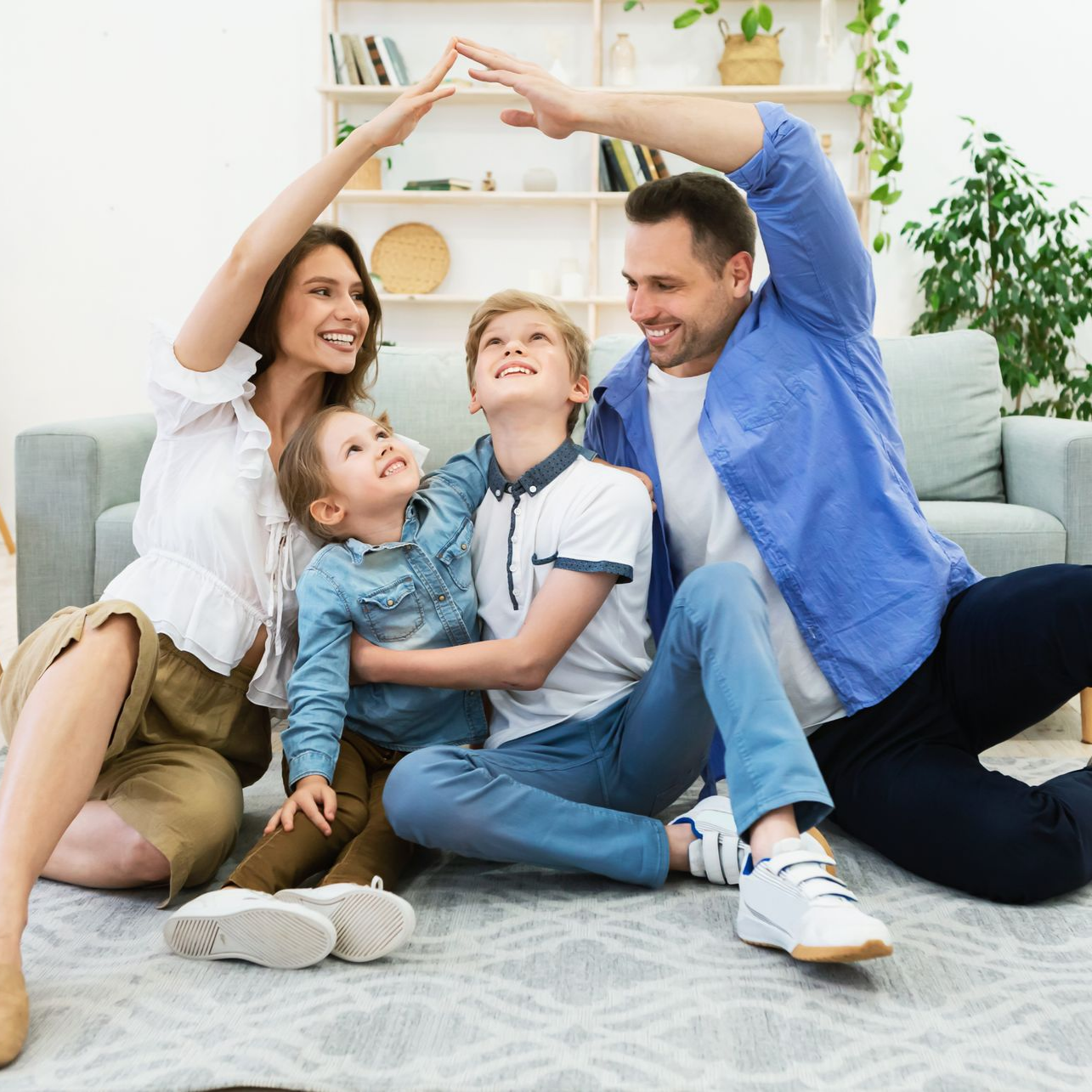Une famille assise sur un tapis de salon, souriante et formant un toit avec ses bras.