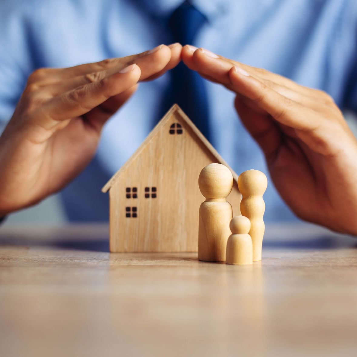 Des mains protégeant une petite maison en bois et deux figurines sur une table.