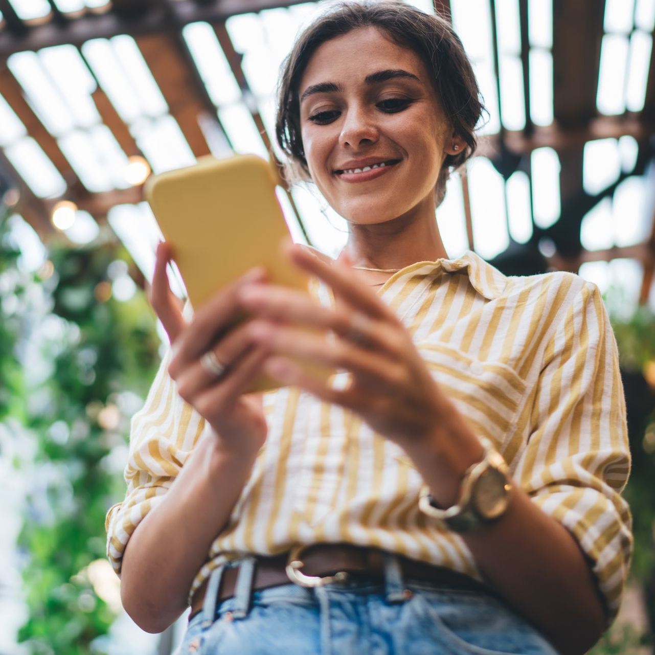 Une personne souriante regarde son téléphone à l'extérieur, sous une pergola en bois.