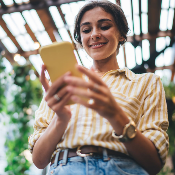 Une personne souriante regarde son téléphone à l'extérieur, sous une pergola en bois.