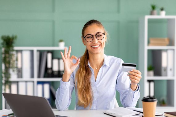 Une femme assise à un bureau, tenant une carte de crédit et faisant un signe OK, dans un bureau lumineux.