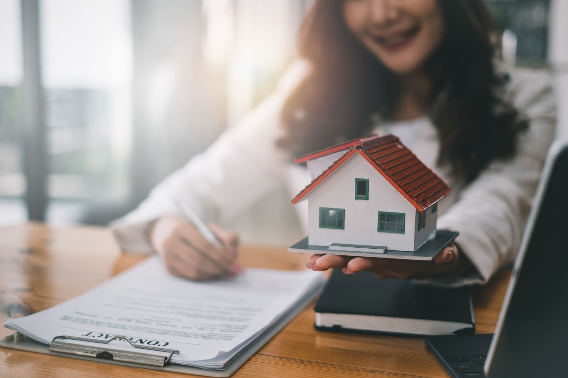 Une personne signe des documents à côté d'une maquette de maison posée sur un bureau, un ordinateur portable à proximité.