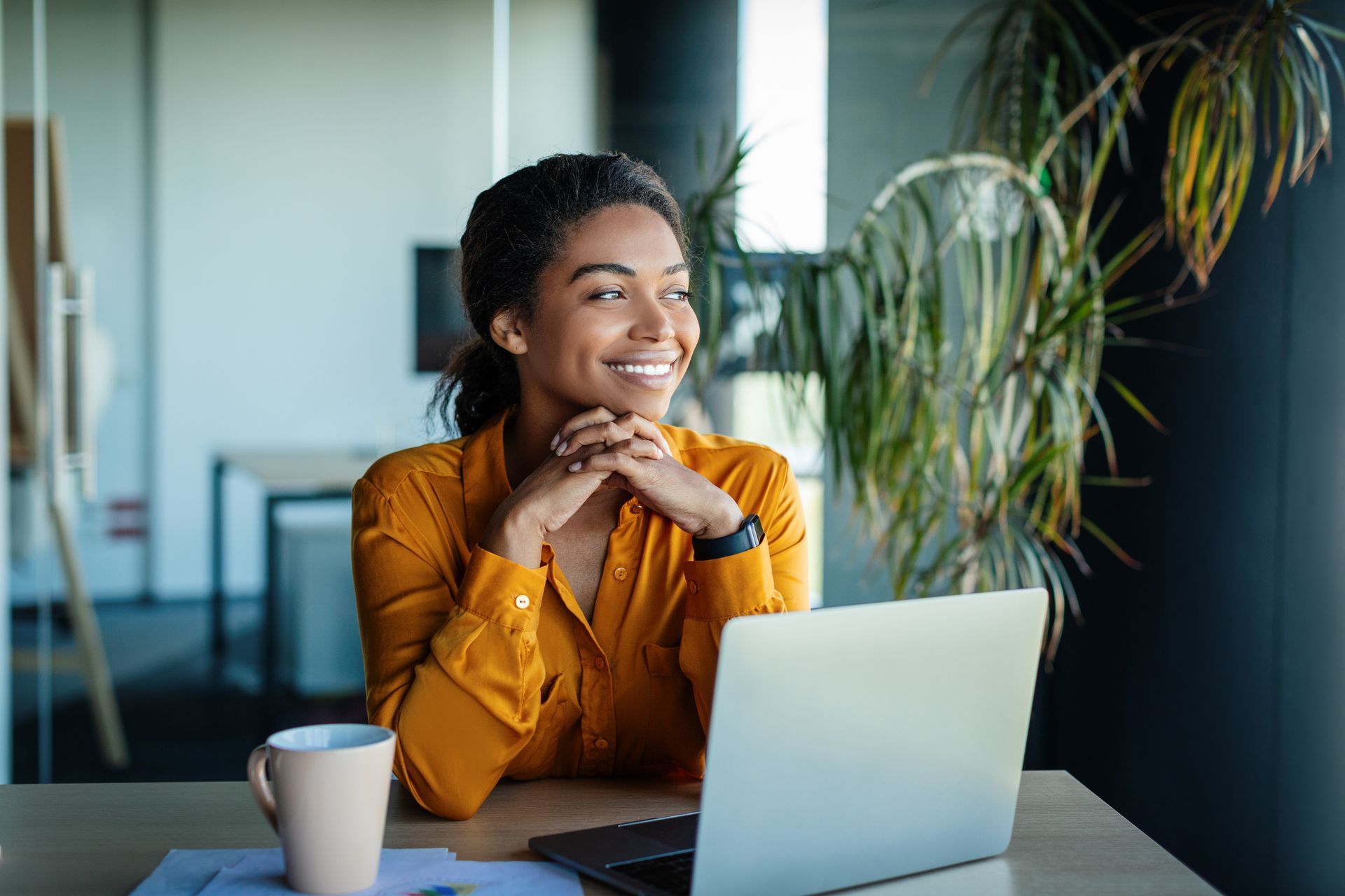 Femme souriante assise à un bureau avec un ordinateur portable et une tasse de café dans un bureau lumineux.