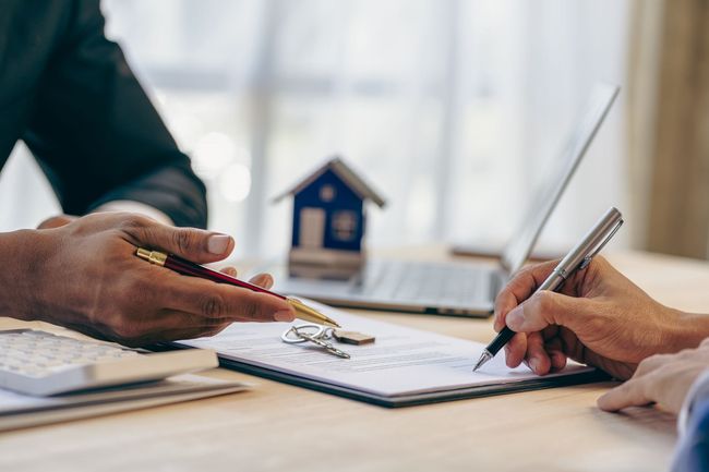Des mains signent des documents à un bureau, avec une maquette de maison en arrière-plan.