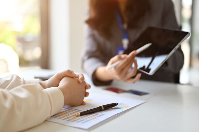 Une personne tient une tablette tandis qu'une autre pose ses mains jointes sur une table où se trouvent des documents et un stylo.
