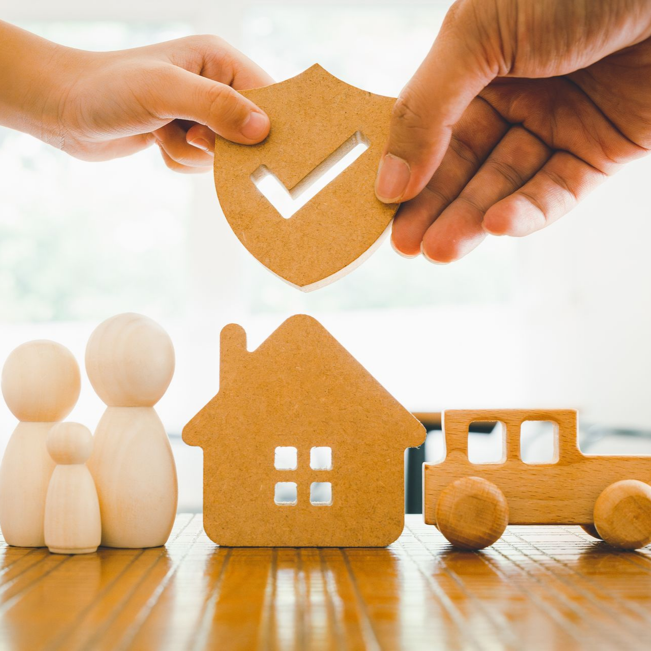 Des mains placent un bouclier beige avec une coche sur une maison de jouets, des figurines familiales et une voiture posées sur une table en bois.