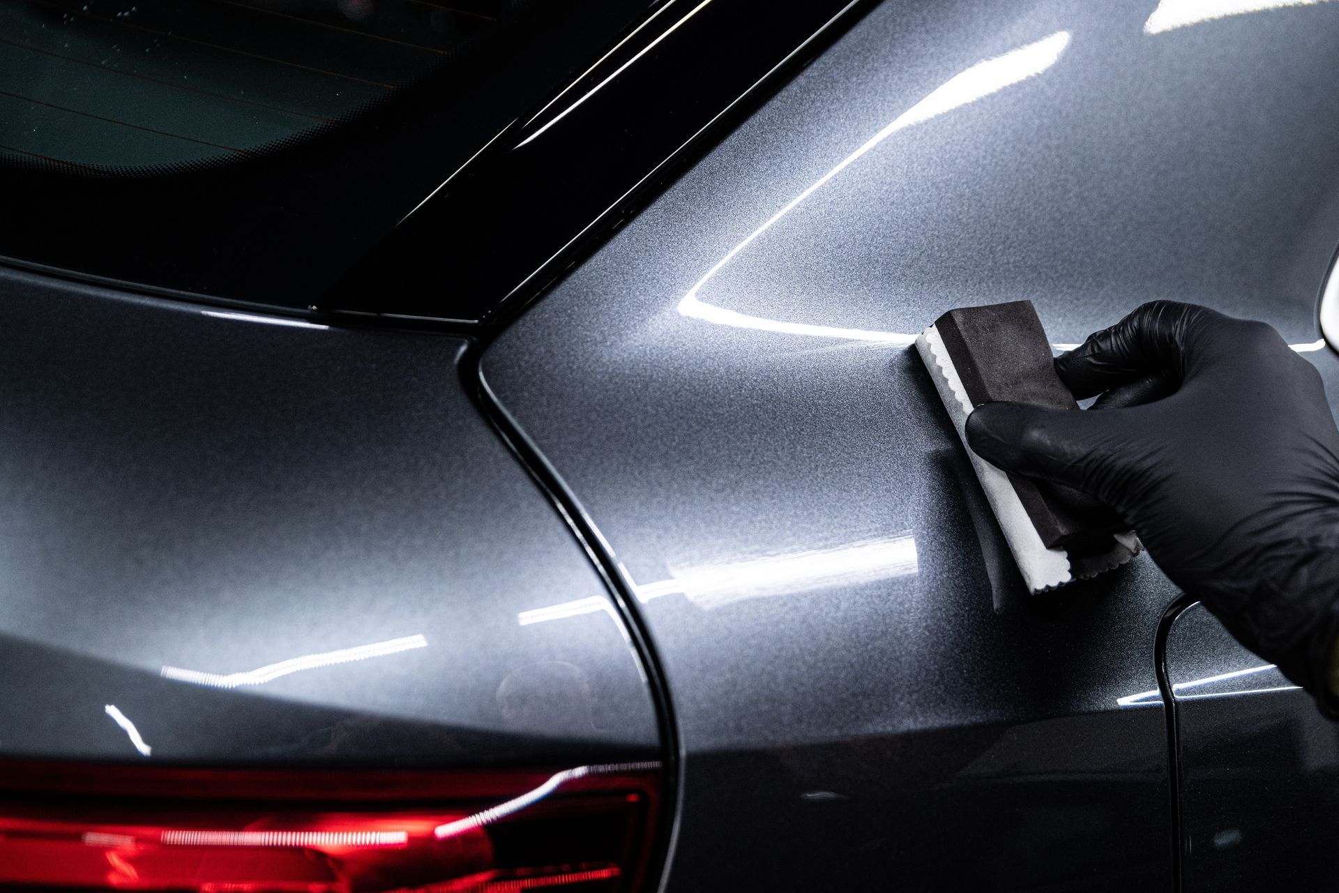 Behandschuhte Hand trägt eine Keramikbeschichtung auf ein dunkelgraues Autoblech auf und verstärkt so seinen Glanz.