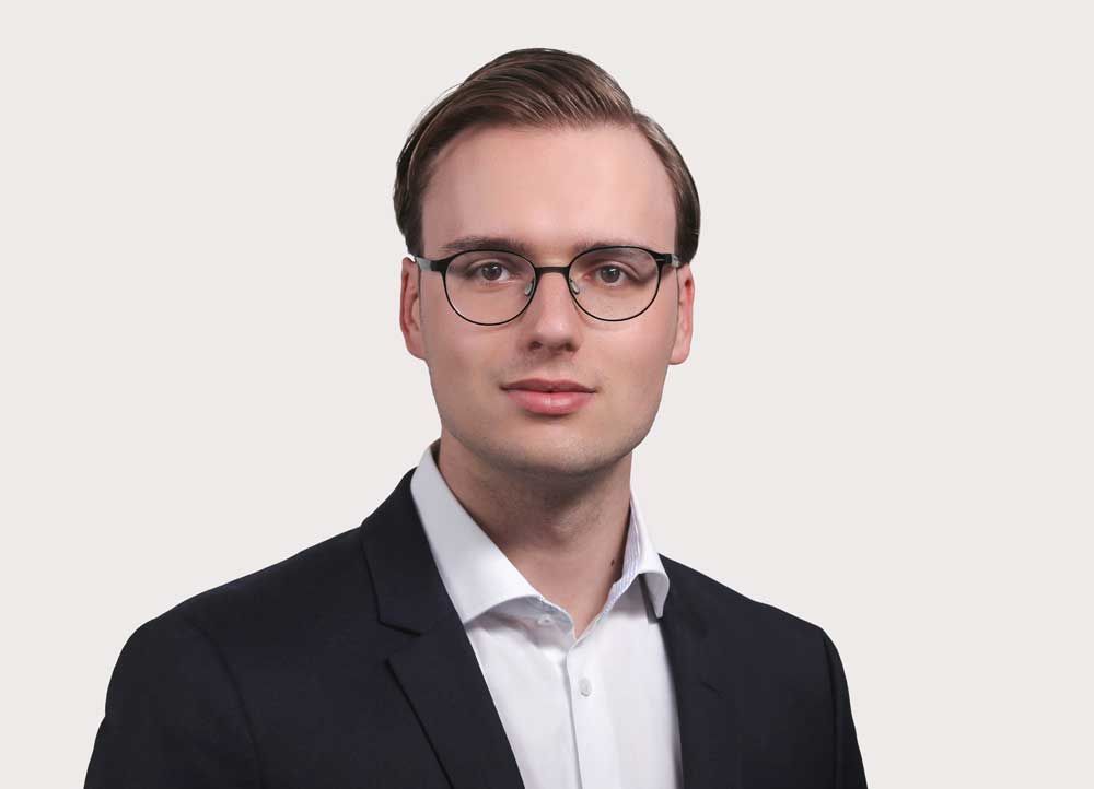 Ein junger Mann mit Brille und Anzug steht vor einem weißen Hintergrund.