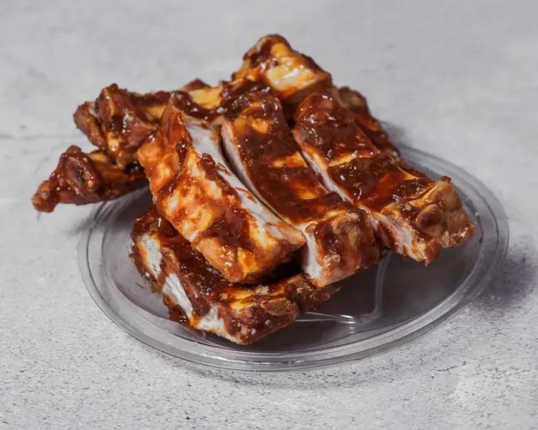 Costillas glaseadas brillantes apiladas en un pequeño plato transparente sobre un fondo gris.
