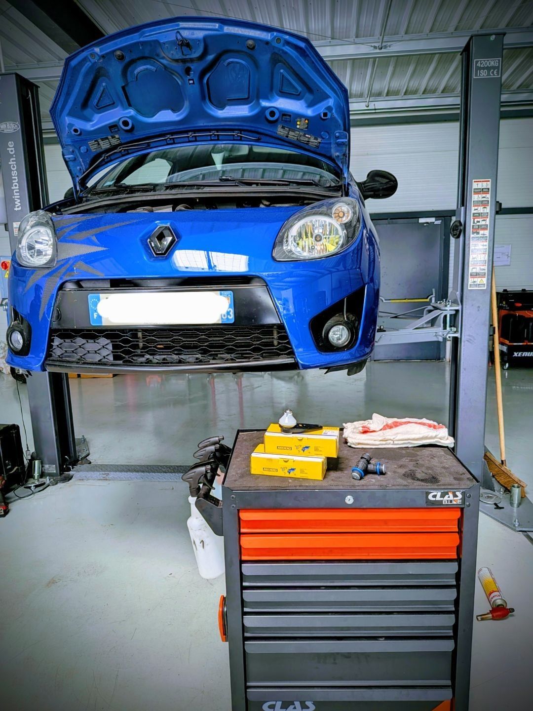 Voiture bleue soulevée dans un garage pour réparation, capot ouvert et caisse à outils devant.