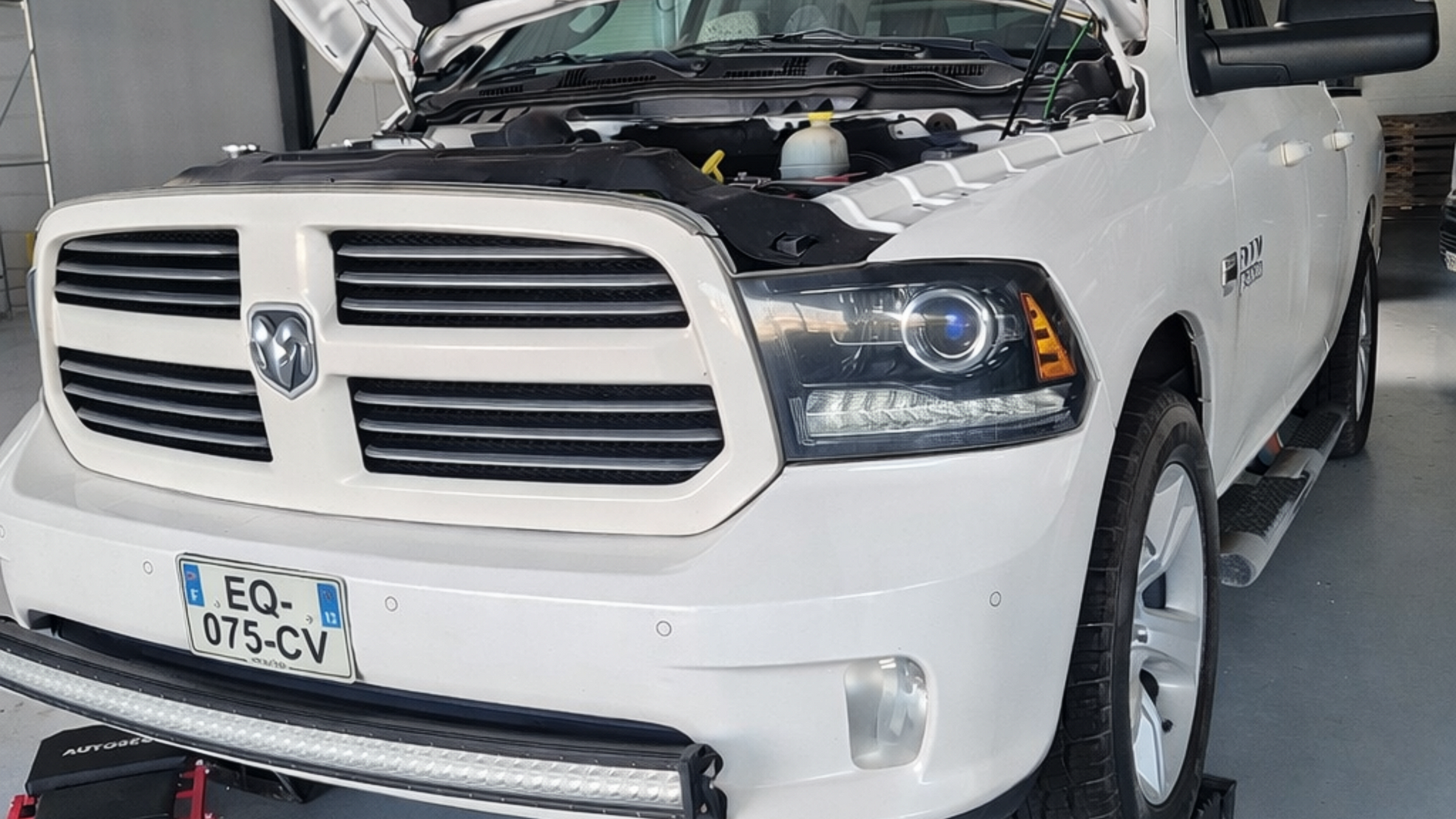 Un pick-up Dodge Ram blanc, capot ouvert, garé dans un garage, avec une barre lumineuse proéminente sur le pare-chocs avant.