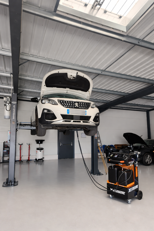Une voiture blanche est soulevée sur un pont élévateur dans un garage, avec un chargeur de batterie orange en dessous.