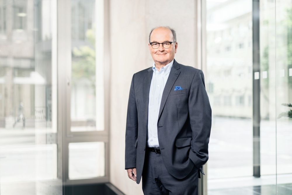 Ein lächelnder Geschäftsmann in dunklem Blazer und weißem Oberteil steht an einem Fenster in einem hellen, modernen Innenraum.