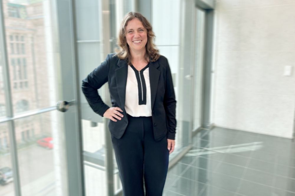 Eine Person in einem dunkelblauen Anzug und einem hellen Hemd lächelt und steht in einem hellen Büroflur mit bodentiefen Fenstern.