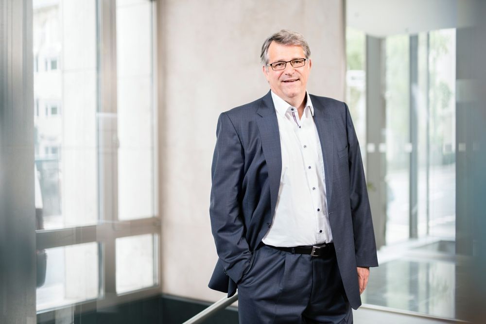 Eine Person im Anzug und mit Brille lächelt in einem hellen Büroflur.