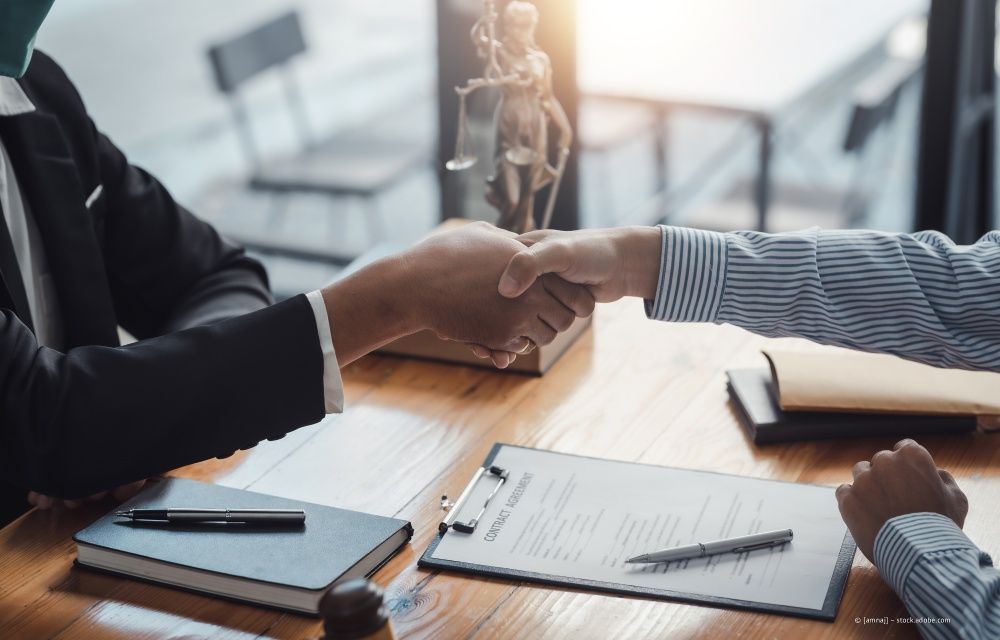 Zwei Personen schütteln sich in einem Büro mit einer Justitia-Statue die Hände über einem juristischen Dokument und einem Notizbuch.