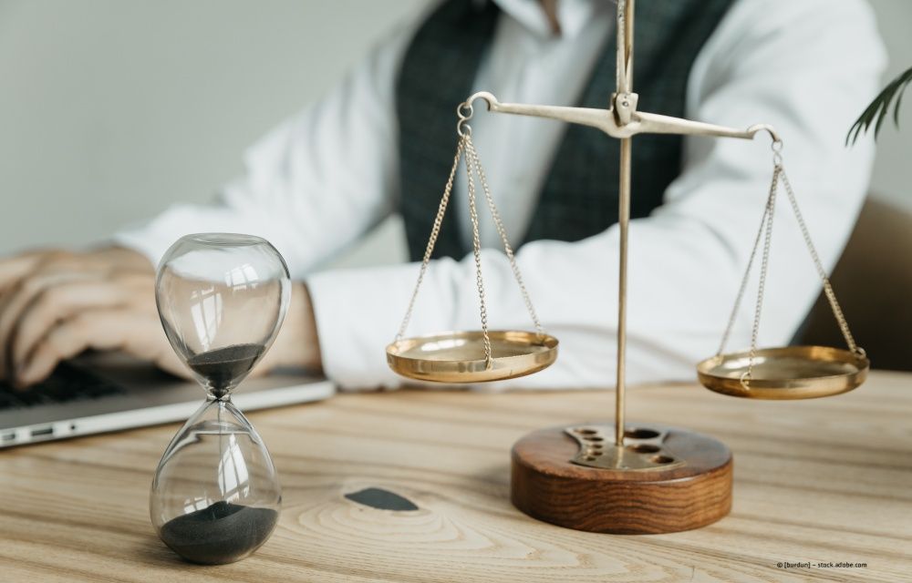 Eine Person in professioneller Kleidung arbeitet an einem Schreibtisch neben einer Sanduhr und einer goldenen Waage der Gerechtigkeit.