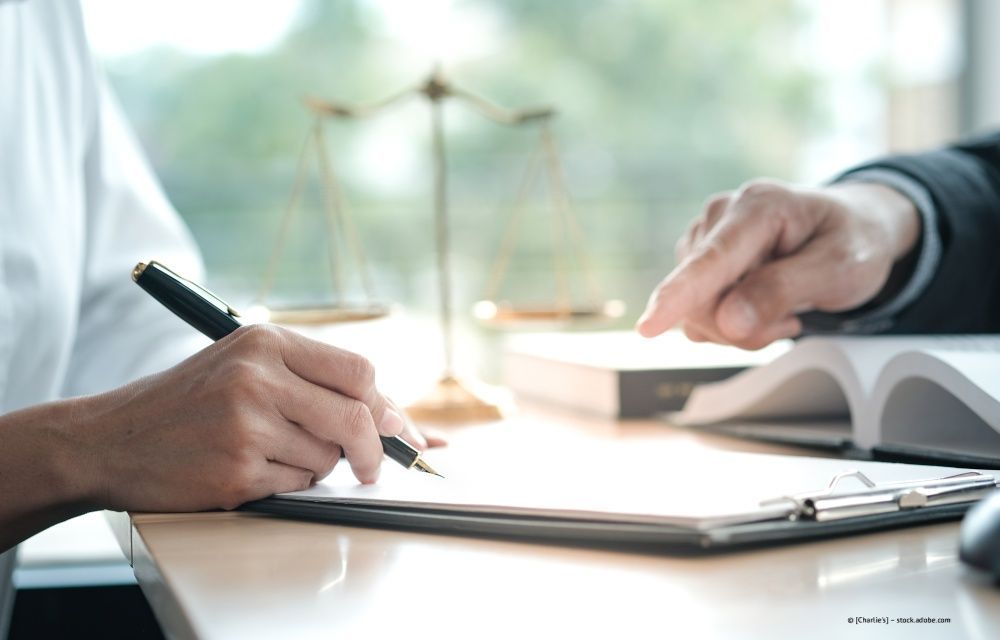 Eine Person unterzeichnet ein Dokument an einem Schreibtisch, während ein Anwalt auf eine Klausel in der Nähe einer Justitia-Waage zeigt.