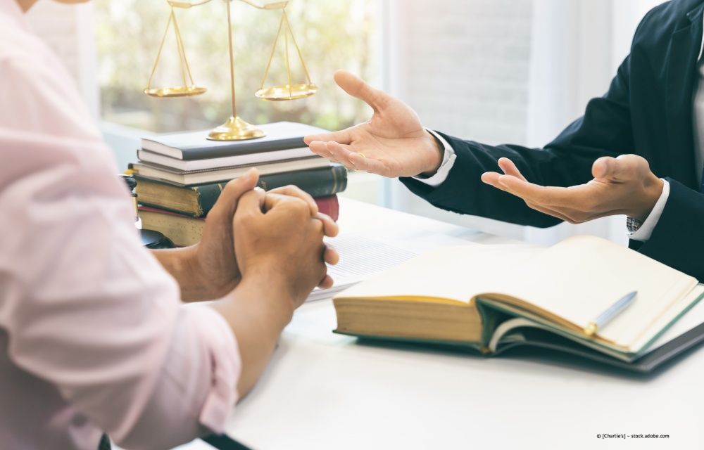 Ein Anwalt in einem dunklen Anzug gestikuliert, während er mit einem Mandanten über einen Schreibtisch hinweg spricht, auf dem Gesetzbücher und eine Justitiawaage stehen.