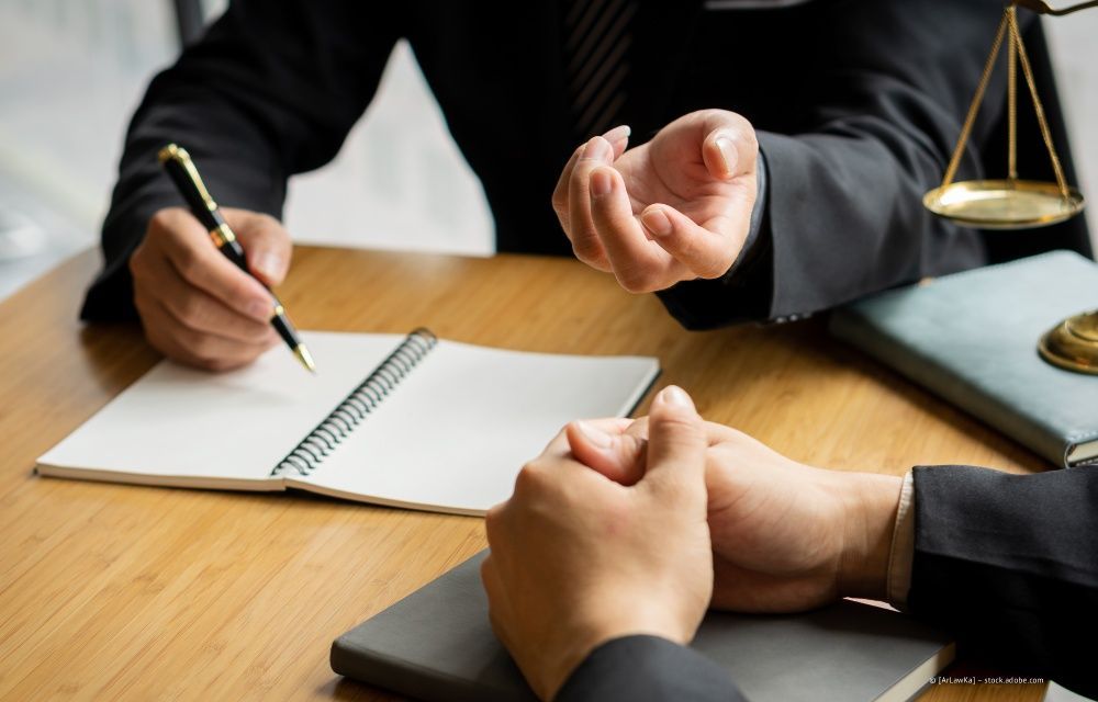 Eine Person im Anzug macht sich Notizen, während eine andere Person ihr gegenüber an einem Holzschreibtisch sitzt, neben dem sich eine Justitiawaage befindet.