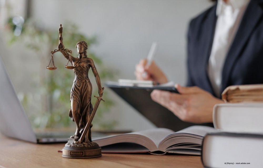 Eine bronzene Justitia-Statue auf einem Schreibtisch mit einem aufgeschlagenen Buch, einem Laptop und einer Person, die beruflich auf einem Klemmbrett schreibt.