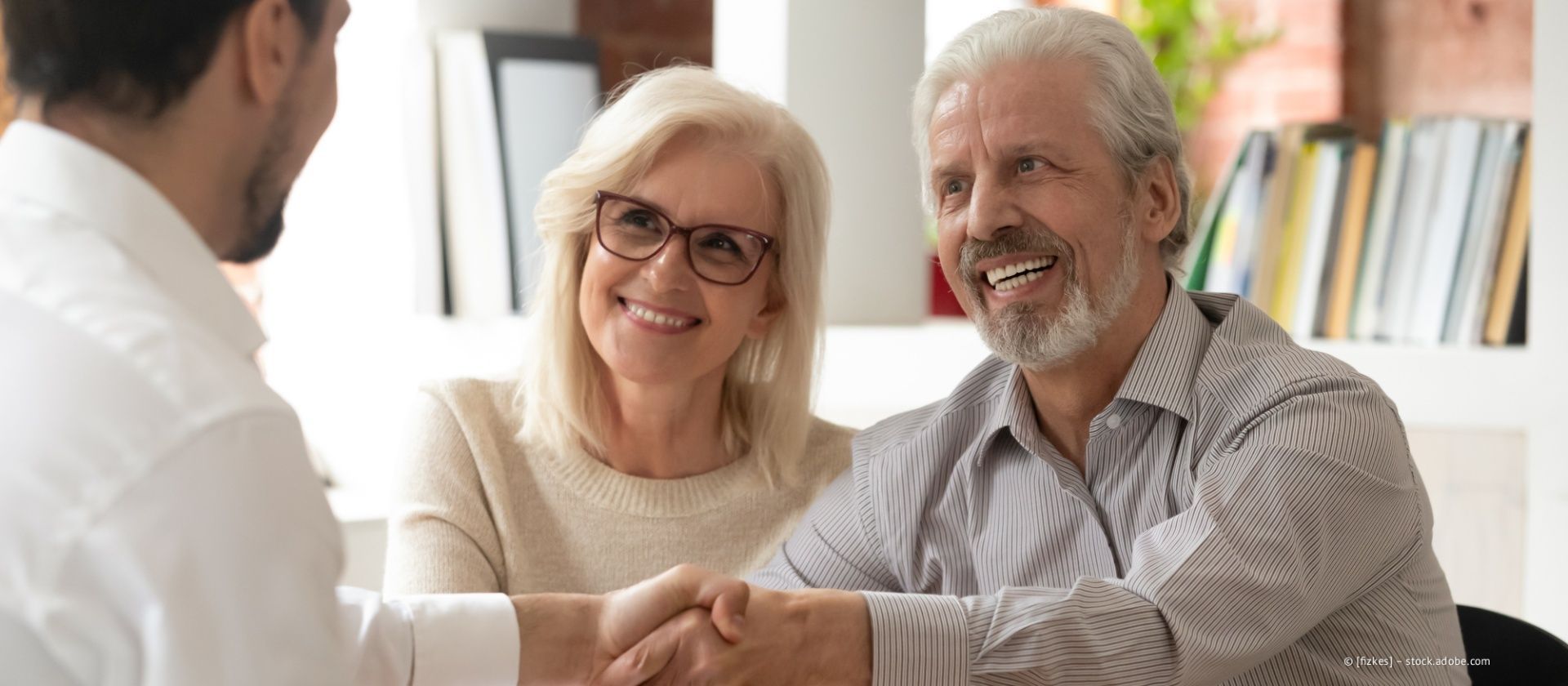 Eine Person schüttelt einem lächelnden Paar die Hand in einem hellen, büroähnlichen Raum mit Bücherregalen im Hintergrund.