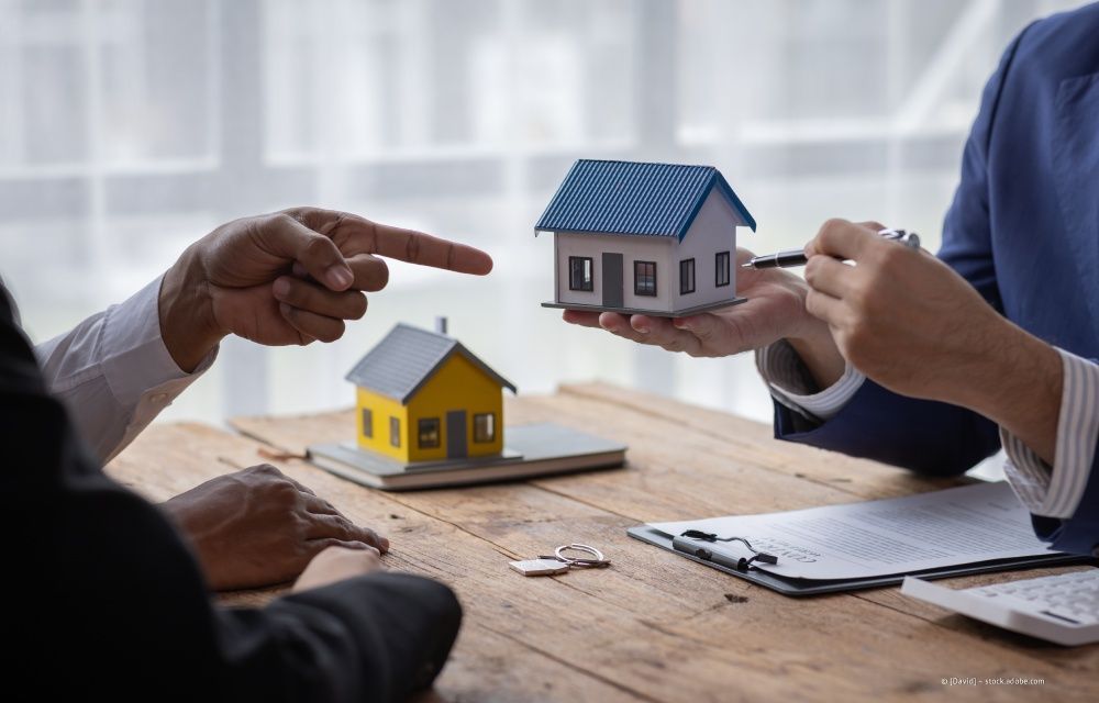 Zwei Personen unterhalten sich auf einem Holztisch anhand von Miniaturhausmodellen und Dokumenten über Immobilien.