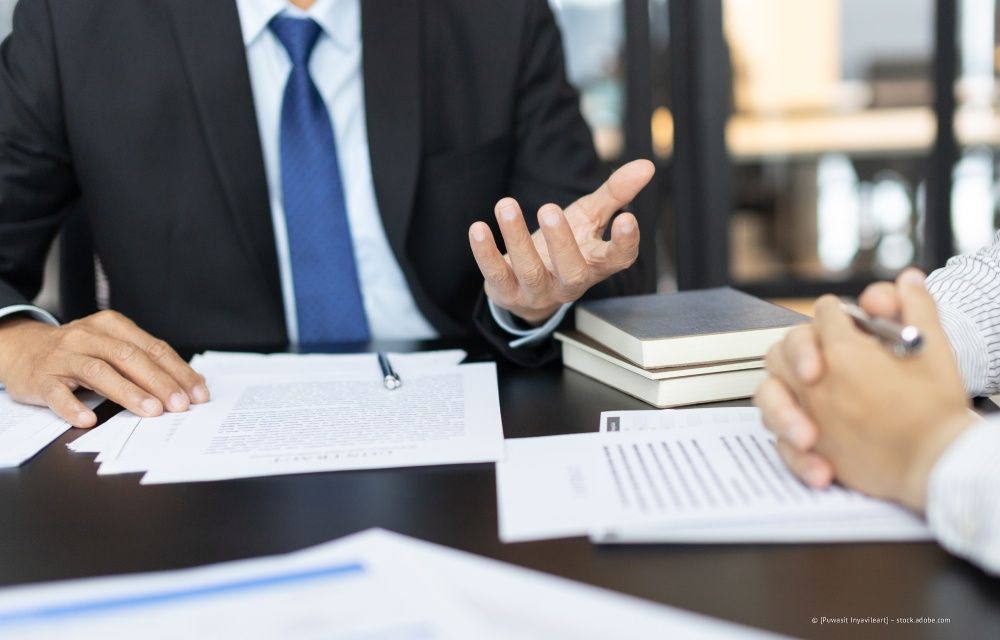 Zwei Personen in Geschäftskleidung sitzen an einem Tisch und gehen Dokumente durch, wobei eine Person beim Sprechen gestikuliert.