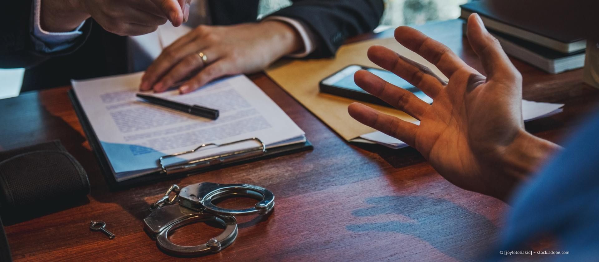 Auf einem Schreibtisch liegen Handschellen neben juristischen Dokumenten, während sich zwei Personen unterhalten, wobei eine mit offener Hand gestikuliert.