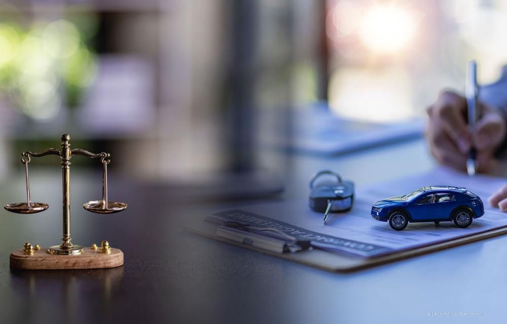 Eine goldene Justitiawaage, ein blaues Spielzeugauto und Schlüssel liegen auf einem Dokument, während im Hintergrund eine Person Papierkram unterschreibt.