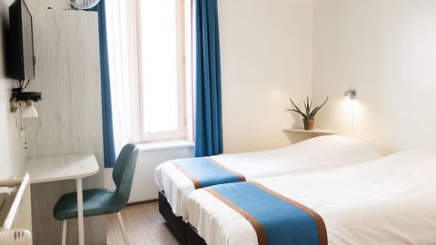 Small hotel room with two twin beds, blue curtains, a desk, and a window.