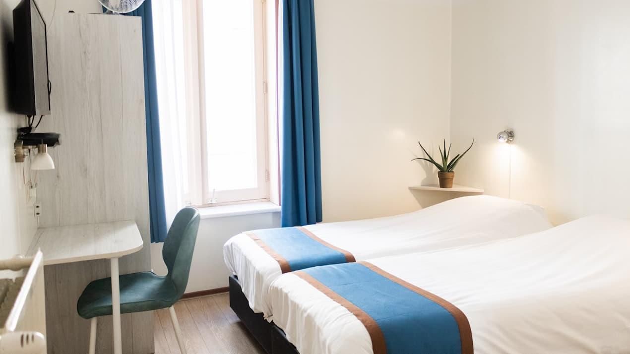 Bright hotel room with twin beds, blue accents, a desk, and a window with curtains