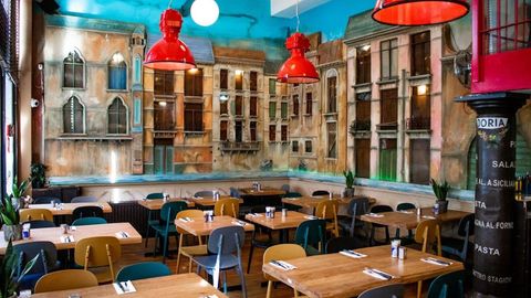 Empty restaurant dining room with wooden tables, black chairs, red pendant lights, and a mural of a city street