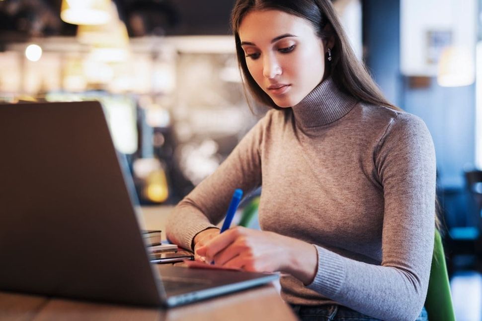 Una mujer está sentada en una mesa usando una computadora portátil y escribiendo en un trozo de papel.