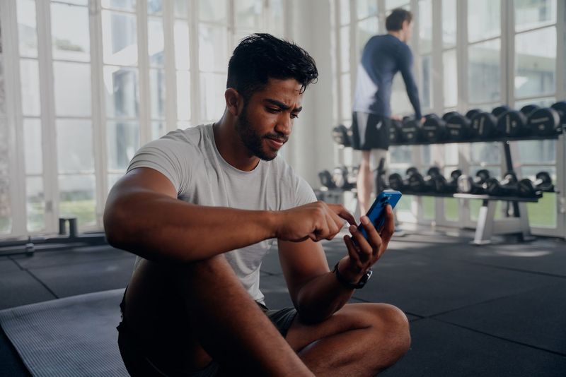 Un hombre está sentado en el suelo en un gimnasio mirando su teléfono celular.
