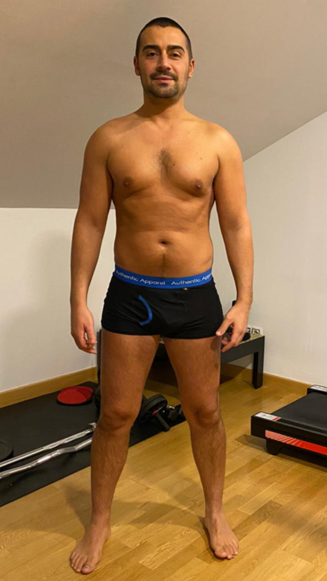 Un hombre sin camisa, con ropa interior negra, está de pie en una habitación.