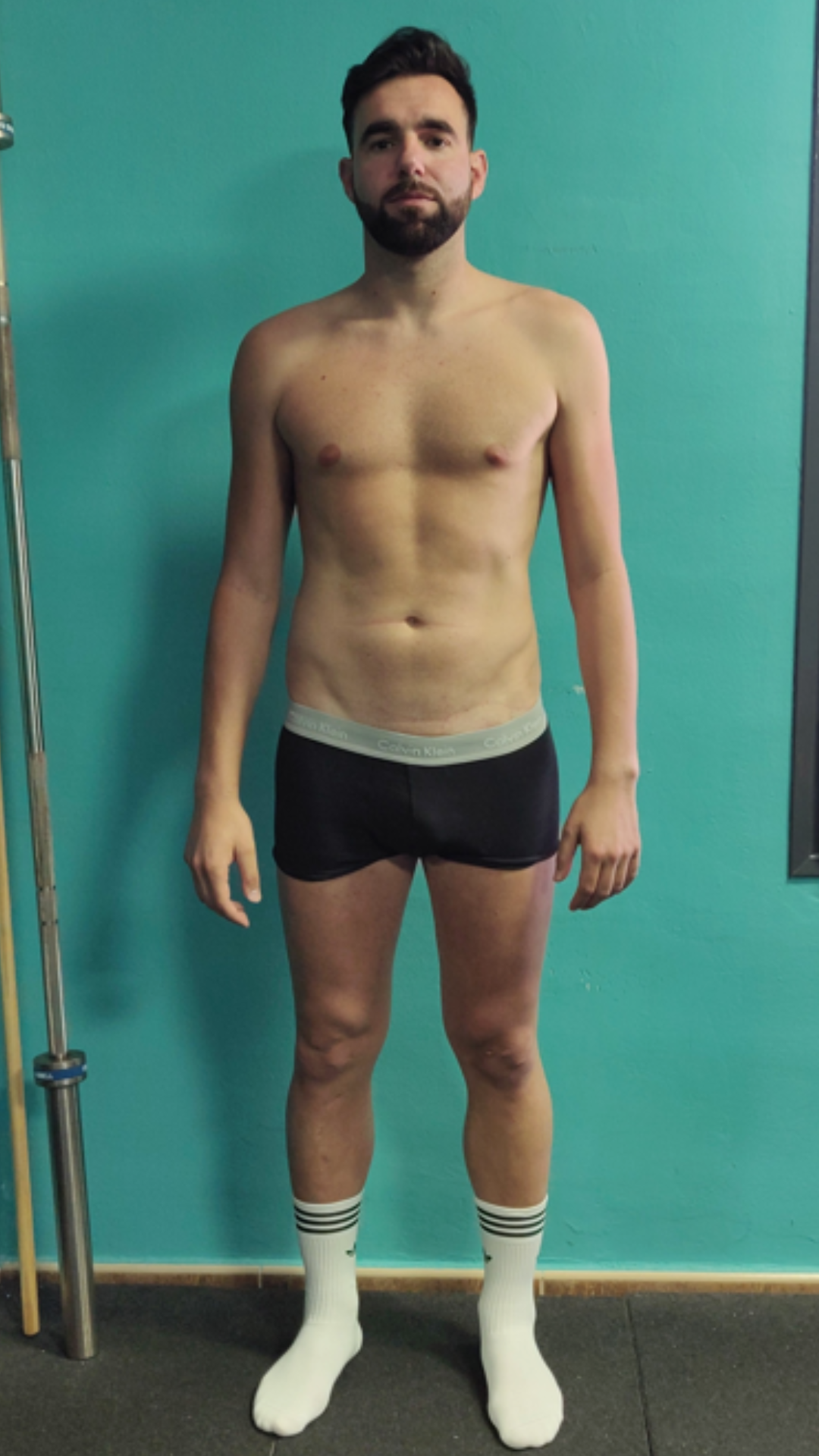 Un hombre sin camisa, con ropa interior negra y calcetines blancos, está parado frente a una pared azul.