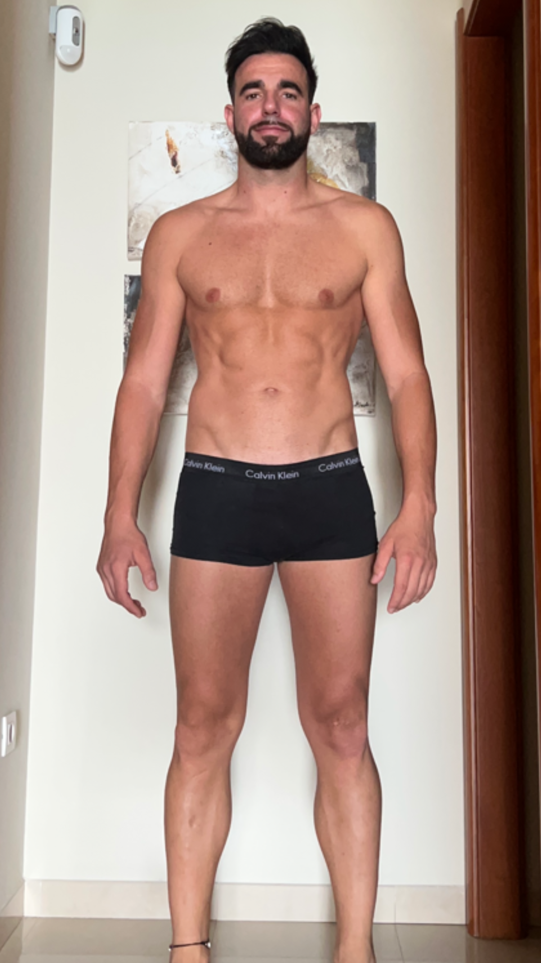Un hombre sin camisa, con ropa interior negra, está de pie en una habitación.