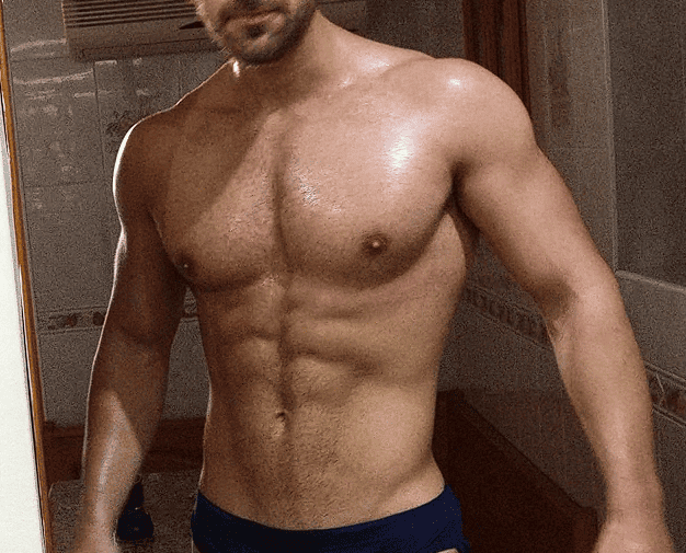 Un individuo en buena forma física, sin camisa, se encuentra de pie en un baño, mostrando un torso musculoso y abdominales definidos.