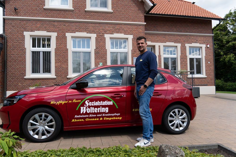 Ein Mann steht neben einem roten Markenauto, das vor einem Backsteinhaus geparkt ist.