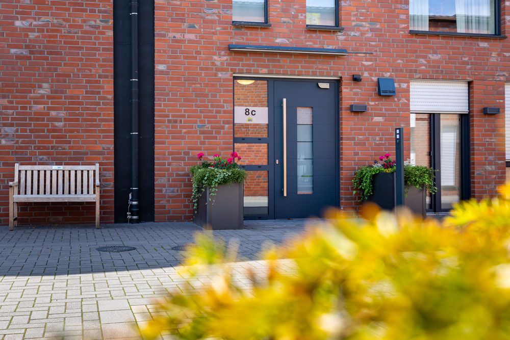 Eingang zum Apartmentgebäude aus Backstein mit grauer Tür, Blumenkästen und Bank an einem gepflasterten Gehweg.