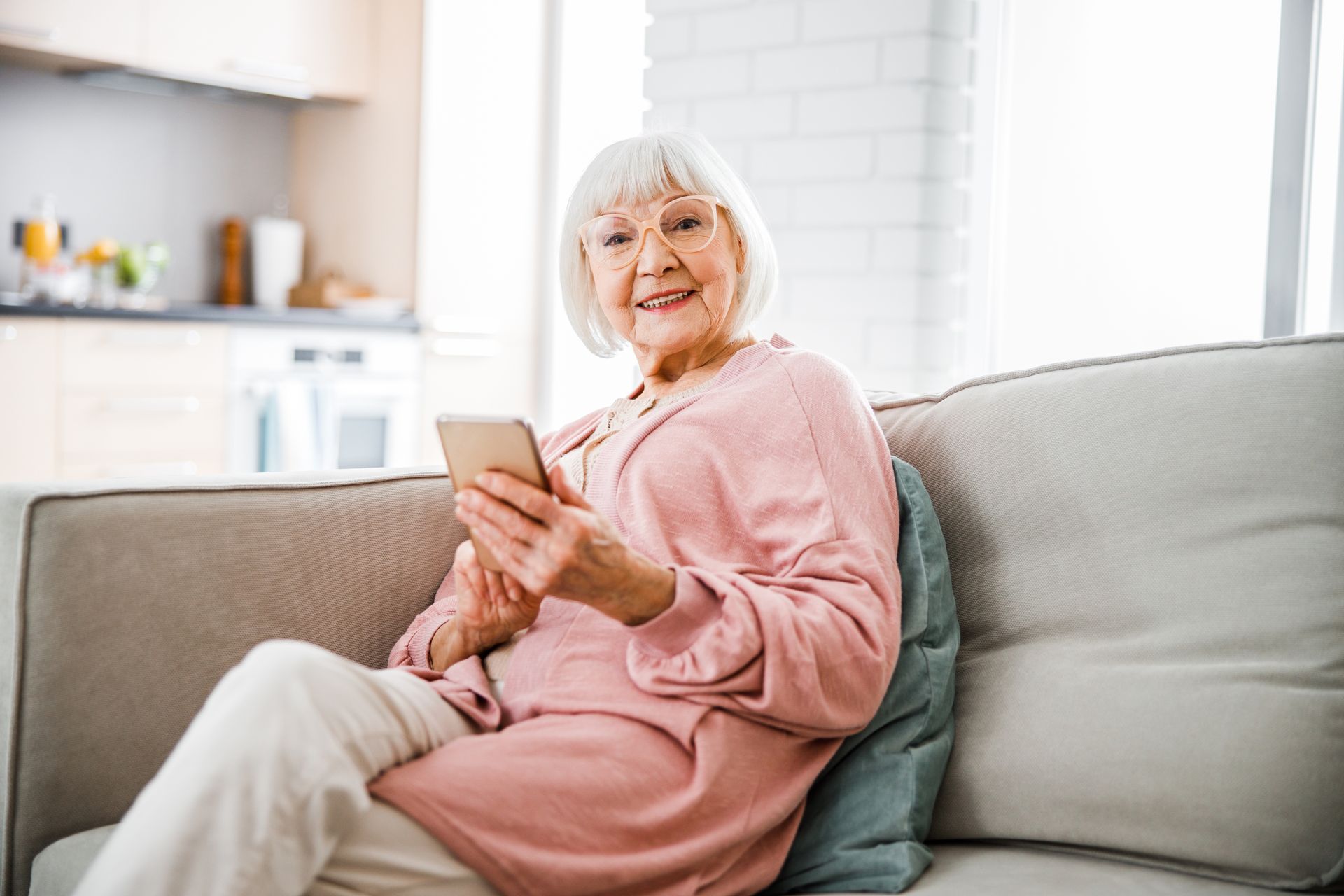 Eine lächelnde ältere Frau in einem rosa Outfit sitzt in einem hellen Wohnzimmer auf einem Sofa und hält ein Telefon in der Hand.