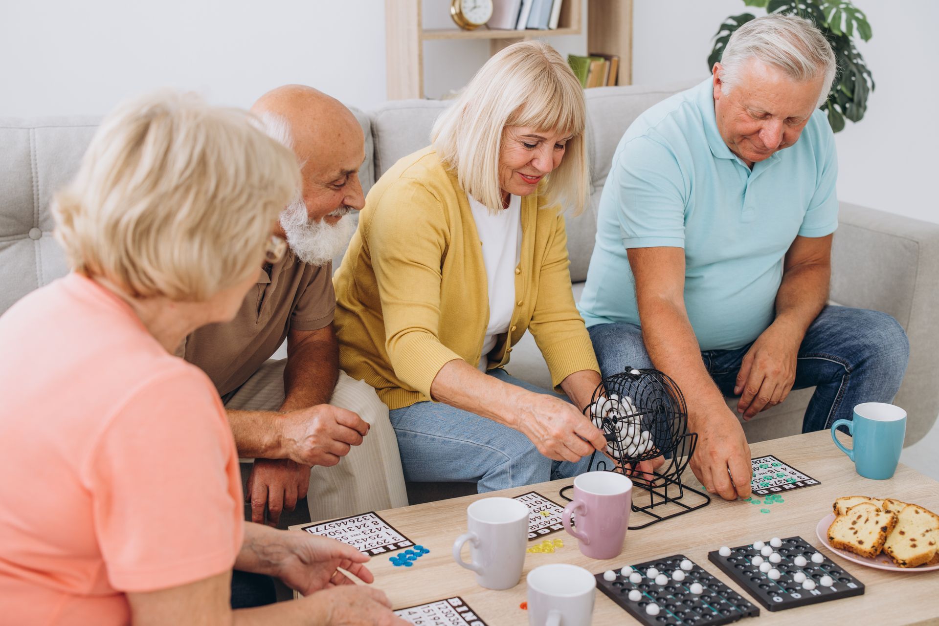 Vier Erwachsene spielen an einem Tisch ein Brettspiel und greifen nach den Spielsteinen; Getränke und Snacks stehen in der Nähe.