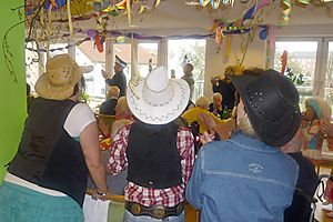 Menschen sitzen bei einer festlichen Zusammenkunft in einem Innenraum, von hinten gesehen, mit Cowboyhüten und bunten Luftschlangen über ihnen.