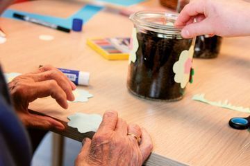 Hände verzieren ein Glasgefäß mit Papierfiguren an einem Basteltisch