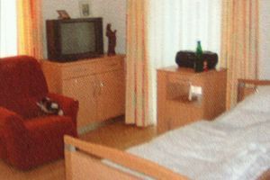 Kleines Schlafzimmer mit Bett, rotem Sessel, Fernseher auf einer Kommode und Vorhängen am Fenster.