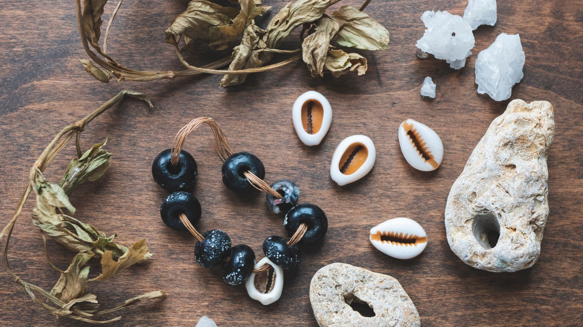Plantes séchées, coquillages cauris, perles, cristaux et morceaux de pierre sur une surface en bois.