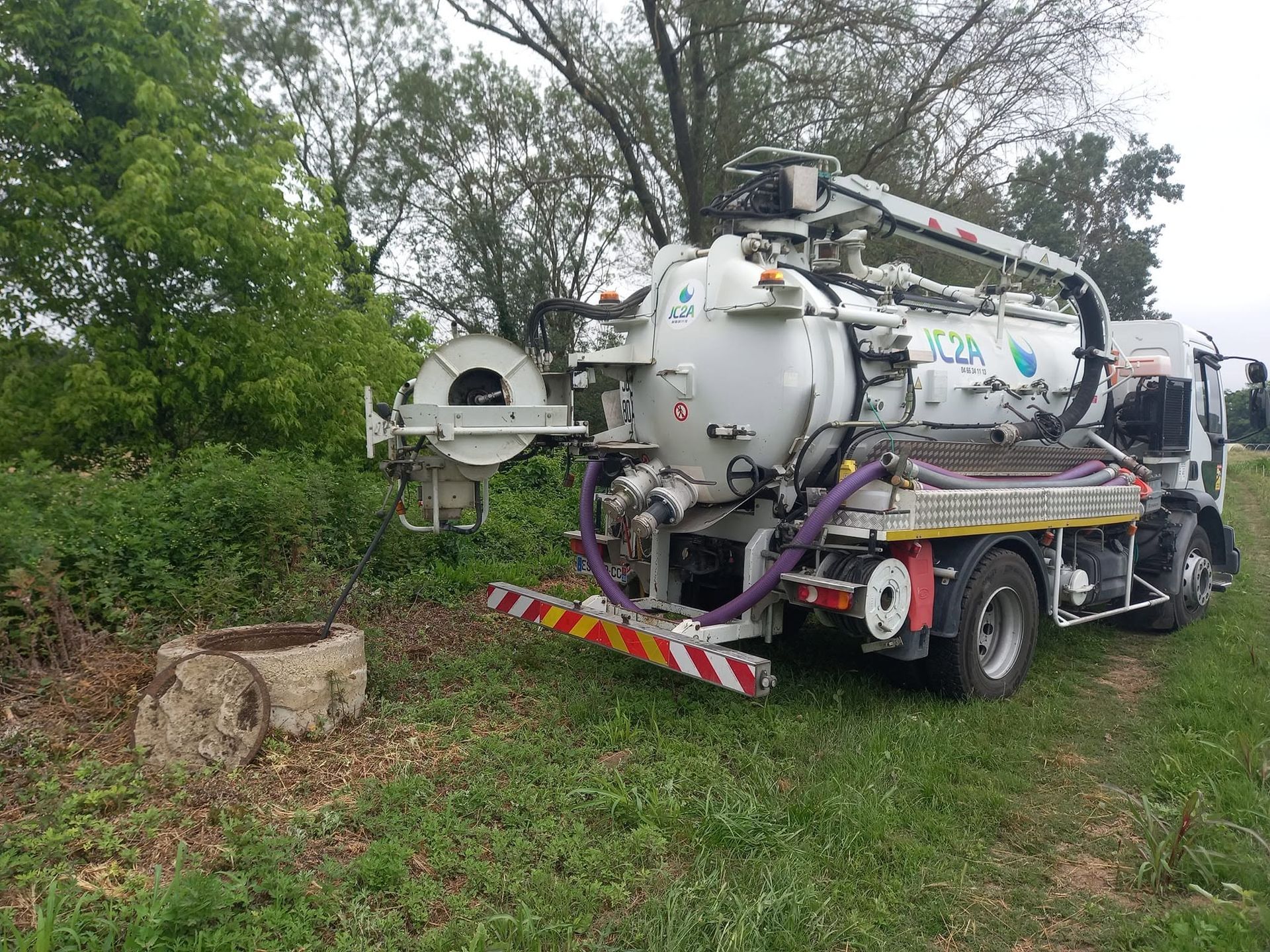Un gros camion aspirateur blanc est stationné sur le bord d'une route herbeuse, à côté d'arbres et de buissons.